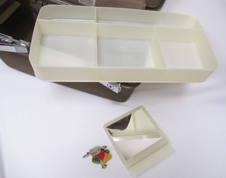May include: A beige plastic organizer tray with multiple compartments, a small mirror, and a key with a colorful charm. The tray is part of a larger brown case, likely for cosmetics or personal items.