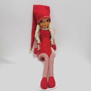 May include: A vintage Christmas elf doll with a red pointed hat, dress, and striped legs. The doll has blonde braids, rosy cheeks, and a small gold bell on the hat. A whimsical holiday decoration.