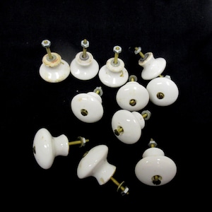 May include: A collection of ten white ceramic cabinet knobs, each featuring a rounded top and a brass-toned metal bolt extending from the base. The knobs include small metal nuts and washers attached to the threaded stems for secure installation.