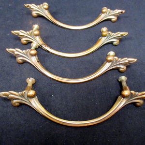 May include: Four antique brass drawer pulls with a curved design. Each pull features decorative leaf and ball accents on the ends. The pulls are arranged on a dark background, showcasing their aged patina and intricate details.