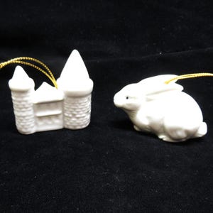 May include: Two white ceramic ornaments: a castle and a rabbit. The castle has two towers and a pointed roof. The rabbit is in a seated position with its ears up. Both ornaments have gold string for hanging.