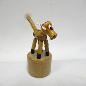 May include: A small wooden animal figurine featuring a tan body with brown spots, black hooves, and a white tufted tail. The creature has white ears with red accents, black eyes, and stands atop a cylindrical wooden base with a smooth finish.