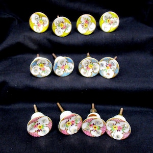 May include: A set of twelve round cabinet knobs featuring a floral pattern with yellow, pink, and white petals, accented by small clear rhinestones. Each knob has a metallic post and a colored lattice border in yellow, blue, or pink hues.