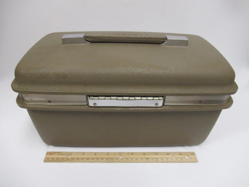 May include: A vintage olive green suitcase with a textured exterior. The suitcase has a silver handle and a silver hinge. A wooden ruler is at the bottom of the image.