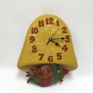 May include: A wall clock shaped like a yellow mushroom with a textured cap. The clock face features raised dark red numbers from 1 to 12. Three black hands indicate the time. The base of the mushroom is brown with green leaves at the bottom.