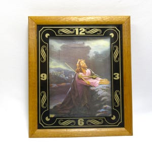 May include: A rectangular wall clock with a light brown wooden frame. The black face features gold numerals 3, 6, 9, and 12 with decorative scrollwork. The center displays a printed image of a figure in a pink robe kneeling in prayer outdoors.