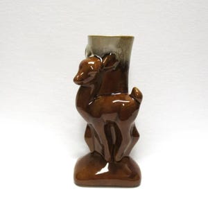 May include: A brown ceramic deer figurine with a glossy finish. The deer is standing next to a tree trunk-shaped vase. The vase has a light brown and beige color. The figurine is on a white background.