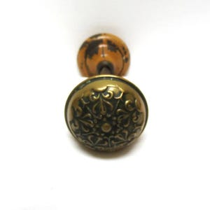 May include: Antique brass door knob with an ornate floral design. The knob has a dark patina and a rounded shape. The back of the knob is a light brown color. The door knob is set against a white background.