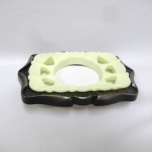 May include: A rectangular belt buckle featuring a dark metallic frame with a textured surface and a central, pale yellow, smooth, molded insert. The insert has a circular opening in the middle and decorative cutouts along its perimeter.