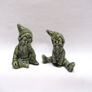 May include: A pair of green ceramic salt and pepper shakers shaped like gnomes. The gnomes have white beards and are sitting with their legs outstretched.