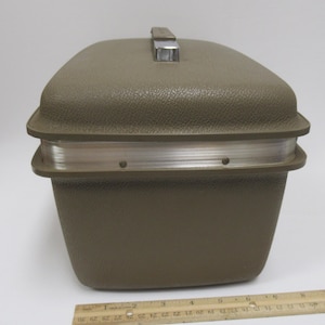 May include: A vintage olive green travel case with a textured exterior and a silver-toned metal handle. The case has a rectangular shape with rounded corners and a silver band around the middle. A wooden ruler is at the bottom.