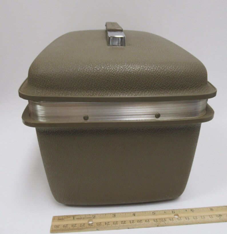 May include: A vintage olive green travel case with a textured exterior and a silver-toned metal handle. The case has a rectangular shape with rounded corners and a silver band around the middle. A wooden ruler is at the bottom.