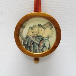 May include: A round, wooden Christmas ornament featuring a painted scene of four children singing carols. The image is framed by a light brown wooden ring and has a small wooden ball at the bottom. A red ribbon is attached for hanging.
