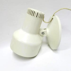 May include: Cream-colored, vintage-style wall-mounted lamp. The lamp features a cylindrical head with vertical slits, a rounded body, and a circular base. A flexible cord extends from the lamp. The lamp is designed to be mounted on a wall.