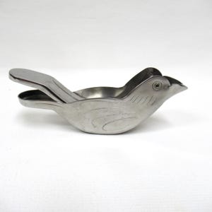 May include: A stainless steel bird-shaped citrus juicer. The juicer has a hinged design with a handle and a spout. The bird's body has etched details, and the juicer is designed to extract juice from citrus fruits.