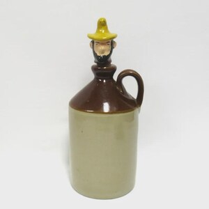 May include: A vintage ceramic jug with a brown top and a tan base. The jug features a handle and a decorative stopper shaped like a man wearing a yellow hat. The stopper has a painted face with a beard. The jug is likely for storing liquids.