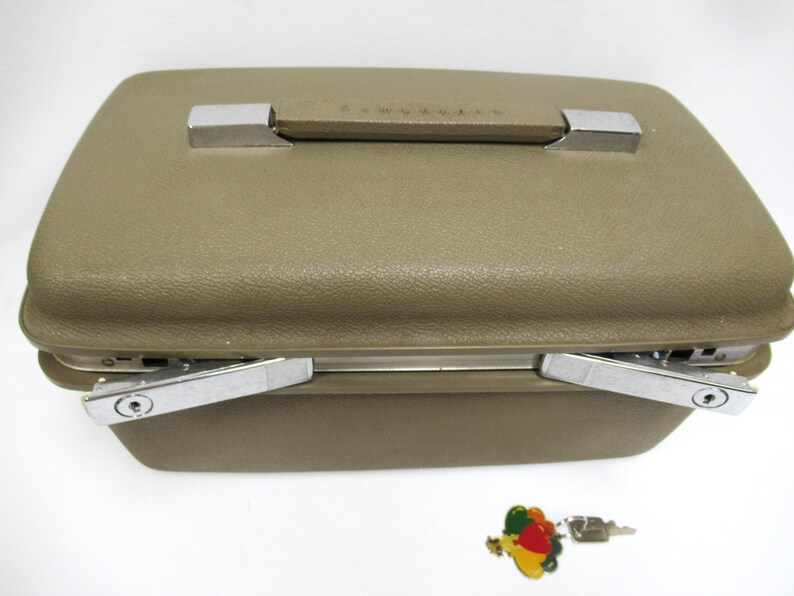 May include: A vintage olive green Samsonite suitcase with a textured surface, chrome accents, and a handle. The suitcase has two chrome latches and a small key with a colorful charm.