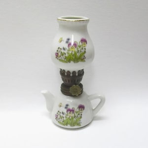 May include: A white ceramic oil lamp with a teapot base and a bulbous top. The lamp is decorated with a floral pattern of purple, yellow, and blue flowers. The lamp has a brown metal base and a gold rim.