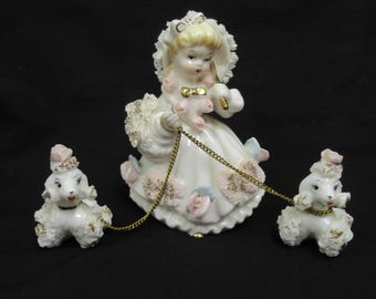 May include: A porcelain figurine of a girl in a white dress and bonnet, holding a gold chain leash attached to two white poodle figurines. The dress is adorned with pink and blue floral accents. The poodles have pink accents.
