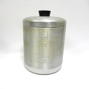 May include: A cylindrical brushed aluminum sugar canister with a vertical rectangular panel featuring the embossed word SUGAR. The canister has a matching removable lid topped with a small, round, black knob handle centered on the top surface.