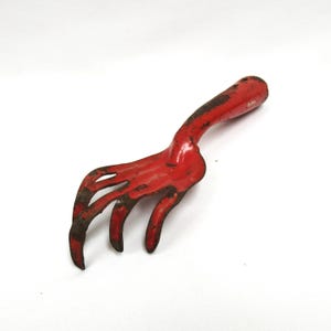 May include: A small, vintage red garden hand rake with a curved handle and four tines. The metal tool shows signs of wear and age, with some rust and paint loss. The rake is designed for small gardening tasks.