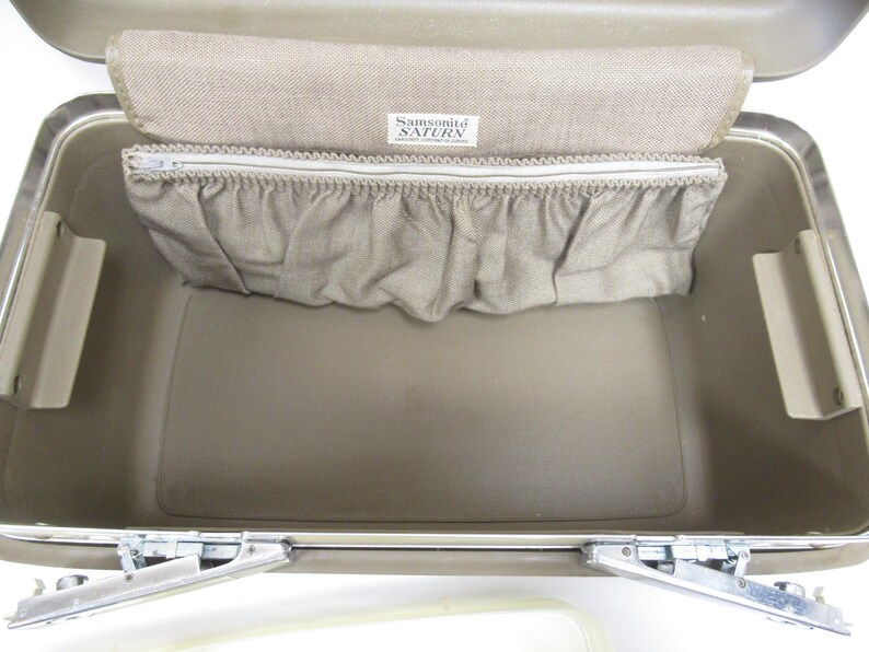 May include: Open view of a vintage Samsonite Saturn suitcase. The interior is a light tan color with a matching fabric pocket and a zippered compartment. The Samsonite Saturn label is visible. The suitcase has metal hinges and clasps.