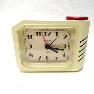 May include: A vintage Jerger quartz alarm clock in cream with a red top button. The clock face has black numerals and hands, with the brand name "Jerger" and "Quartz" printed on the face. The clock is rectangular with a slightly angled face.
