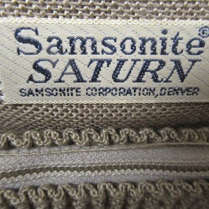 May include: Close-up of a beige fabric label with the text "Samsonite SATURN" in dark blue. Below the name is "SAMSONITE CORPORATION, DENVER". The label is sewn onto a textured, beige fabric.