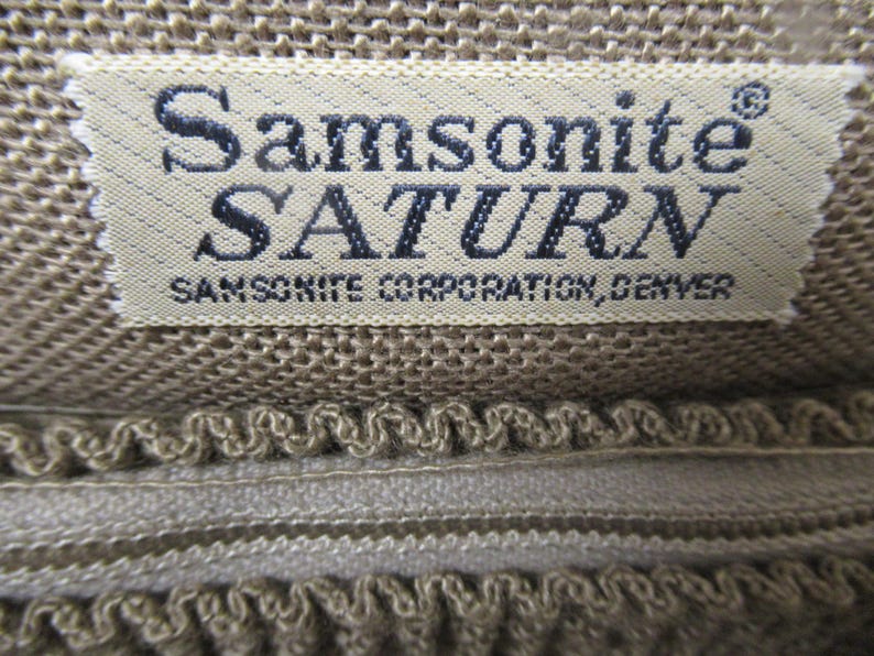 May include: Close-up of a beige fabric label with the text "Samsonite SATURN" in dark blue. Below the name is "SAMSONITE CORPORATION, DENVER". The label is sewn onto a textured, beige fabric.