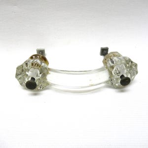 May include: Clear glass cabinet pull with two faceted knobs and a curved handle. The knobs have a brown tint at the top and black screws. The pull is mounted on two small, square, gray metal plates.