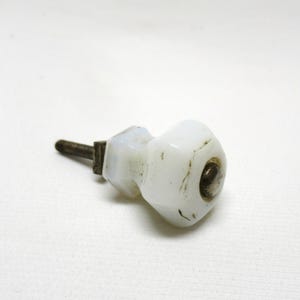 May include: A white, octagonal cabinet knob with a metal stem and a small, dark metal accent. The knob has a slightly aged appearance, with some discoloration. The hardware is likely for a drawer or cabinet.