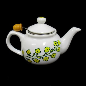 May include: A white ceramic teapot featuring a painted green vine with yellow flowers wrapping around the body. The teapot includes a matching lid with a small knob and a handle attached to a light brown wooden bead on a metal wire extension.