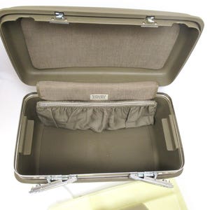 May include: Open olive green vintage suitcase with a tan interior. The suitcase has a rectangular shape with metal latches and hinges. The interior features a fabric-lined top and a zippered pouch with a ruffled edge. The label reads "Samsonite Saturn".