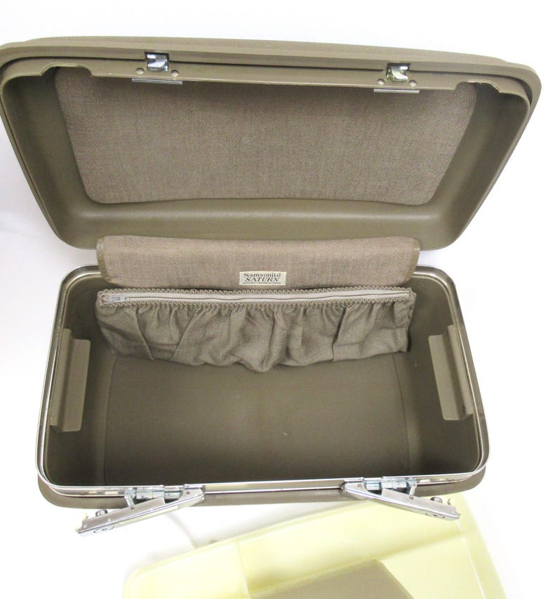 May include: Open olive green vintage suitcase with a tan interior. The suitcase has a rectangular shape with metal latches and hinges. The interior features a fabric-lined top and a zippered pouch with a ruffled edge. The label reads "Samsonite Saturn".