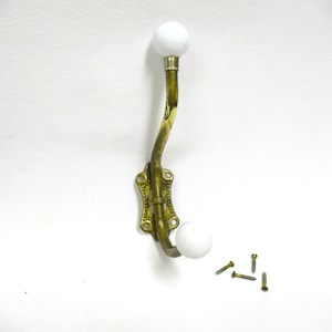 May include: An antique brass wall hook with a curved arm and two white spherical knobs. The hook has a decorative backplate and is accompanied by several small screws, suggesting installation. A vintage-style item.