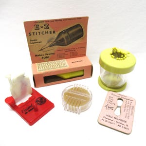 May include: A collection of vintage sewing tools and accessories, including a yellow and white E-Z Stitcher in its original box, a clear plastic pin cushion, a red plastic pin holder with the words "United of Omaha" on it, and a small, clear plastic tool with a yellow interior. The image also includes a pink card with the text "Amazing Scissors Sharpener, Use This Side Only!" and "Hall Transfer, Charleston, Mattoon, Ill. Ph. DI 5-6411 AD 4-3615".