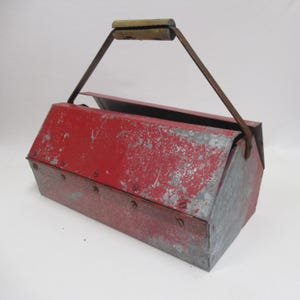 May include: A vintage red metal toolbox with a wooden handle and metal handle supports. The toolbox has a hinged lid and shows signs of wear, with paint chipping and a weathered appearance. The toolbox is designed for carrying tools.