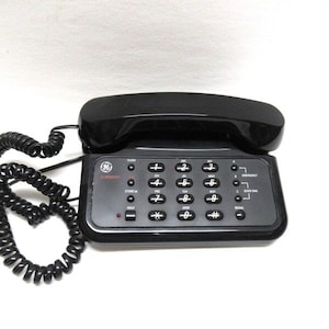 May include: A black corded phone with a rotary dial and a coiled cord. The phone has a black handset and a black base with a keypad and several buttons. The buttons include Flash, Redial, Emergency, Quick Dial, and Hold.