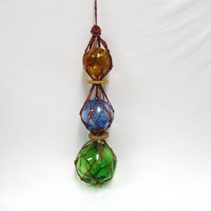 May include: A hanging decorative piece featuring three glass spheres in amber, blue, and green. Each sphere is encased in a brown woven net and separated by wooden rings. The piece is suspended from a brown string.
