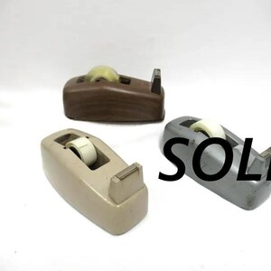 May include: Three desktop tape dispensers in brown, beige, and gray. Each unit features a central roll of clear adhesive tape and a metal cutting blade at the front. The gray dispenser has the word SOLD printed in large black letters across it.