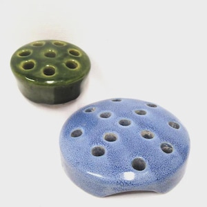 May include: Two ceramic flower frogs. One is a dark green, cylindrical shape with multiple holes. The other is a light blue, rounded shape with a speckled texture and multiple holes. These are used to arrange flowers.