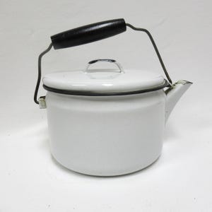 May include: A white enamel tea kettle with a black handle and a matching lid. The kettle features a metal wire handle frame, a small spout on the right side, and a thin black rim around the top edge of the body and the lid. The finish is smooth.