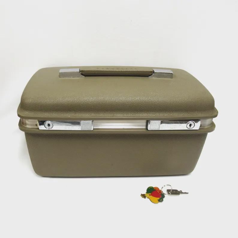May include: A vintage olive green travel case with a silver handle and latches. The case has a rectangular shape and a textured surface. A small, colorful pin and a key are next to the case.