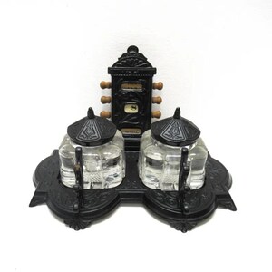 May include: A black cast iron inkwell set with two glass inkwells and a calendar stand. The calendar stand has a wooden knob for turning the date.