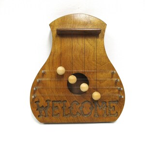 May include: Wooden decorative wall hanging in a musical instrument shape. The instrument is stained a medium brown and features the word "WELCOME" carved into the wood. It has strings and wooden beads. The background is white.