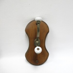 May include: A wall-mounted coat hook featuring a contoured wooden backplate with a medium brown finish. The hook is made of tarnished brass metal and includes two smooth, round white ceramic knobs at the top and bottom for hanging items.