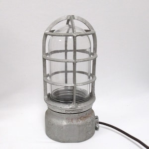 May include: A silver-toned industrial cage light featuring a clear glass dome protected by a metal grid. The base is a heavy, weathered metallic cylinder with a dark electrical cord extending from the side, showcasing a rugged, utilitarian design.