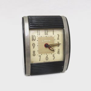 May include: Vintage Westclox alarm clock with a cream-colored face and black numerals. The clock has a rectangular shape with a black, ribbed top and bottom. The clock face features the text "Westclox" and "Made in U.S.A."