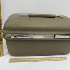 May include: A vintage olive green suitcase with a silver handle and latches. The suitcase is rectangular with rounded edges. A wooden ruler is next to the suitcase. The suitcase measures approximately 16 inches wide.