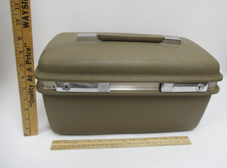 May include: A vintage olive green suitcase with a silver handle and latches. The suitcase is rectangular with rounded edges. A wooden ruler is next to the suitcase. The suitcase measures approximately 16 inches wide.